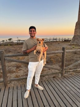 Rapha smiling on a boardwalk at sunset, holding his dog